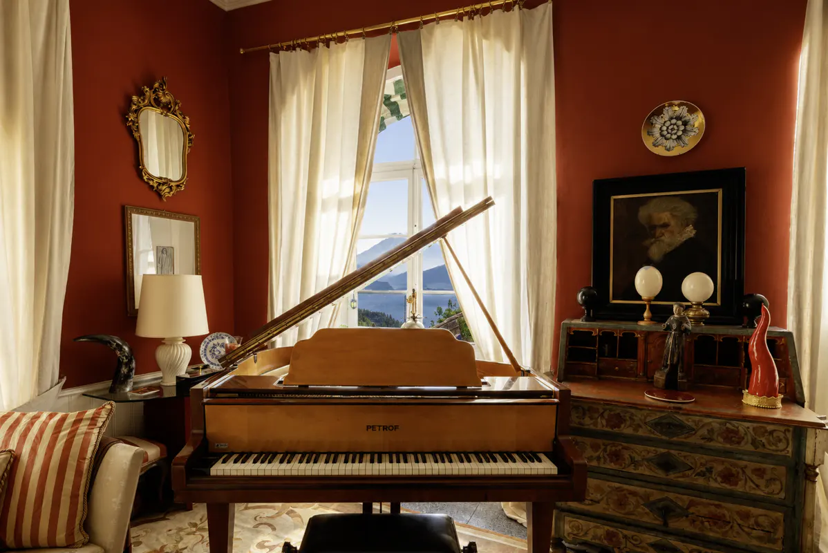 A room with red walls, white curtains, and a light brown grand piano. A painting and ornate mirror decorate the walls.