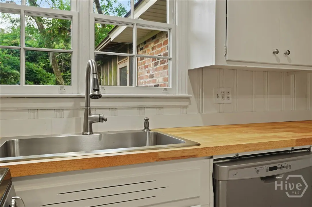 Bright kitchen with stainless steel sink, faucet, and dishwasher. Butcher block countertops and white cabinets. Window view of greenery.