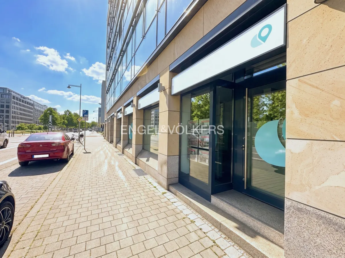 Exterior view of Engel & Völkers office with glass windows and doors on a sunny day. Cars parked on the street.