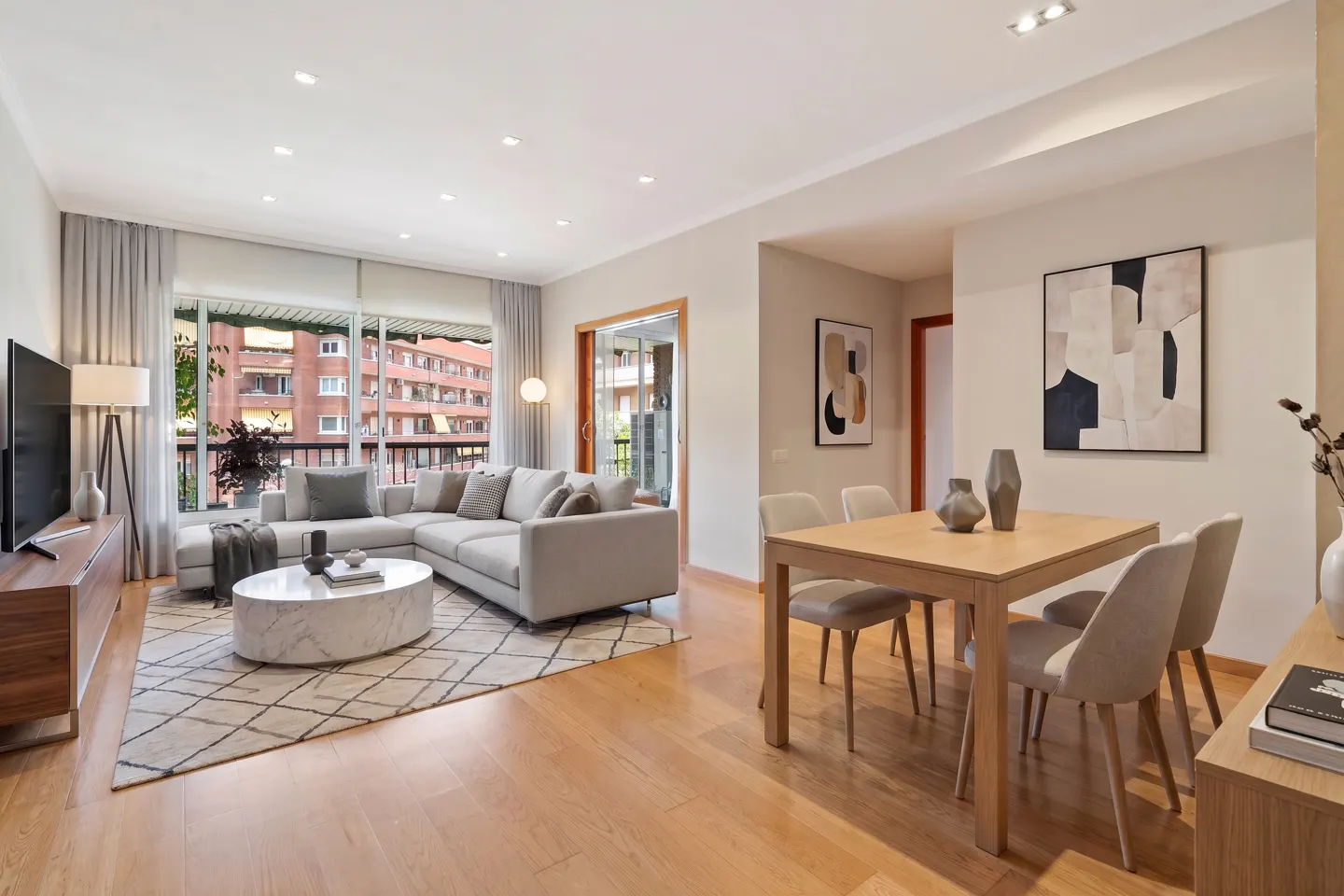 Bright, modern living room with a gray sectional sofa, marble coffee table, and wood dining table with four chairs. Artwork adorns the walls.