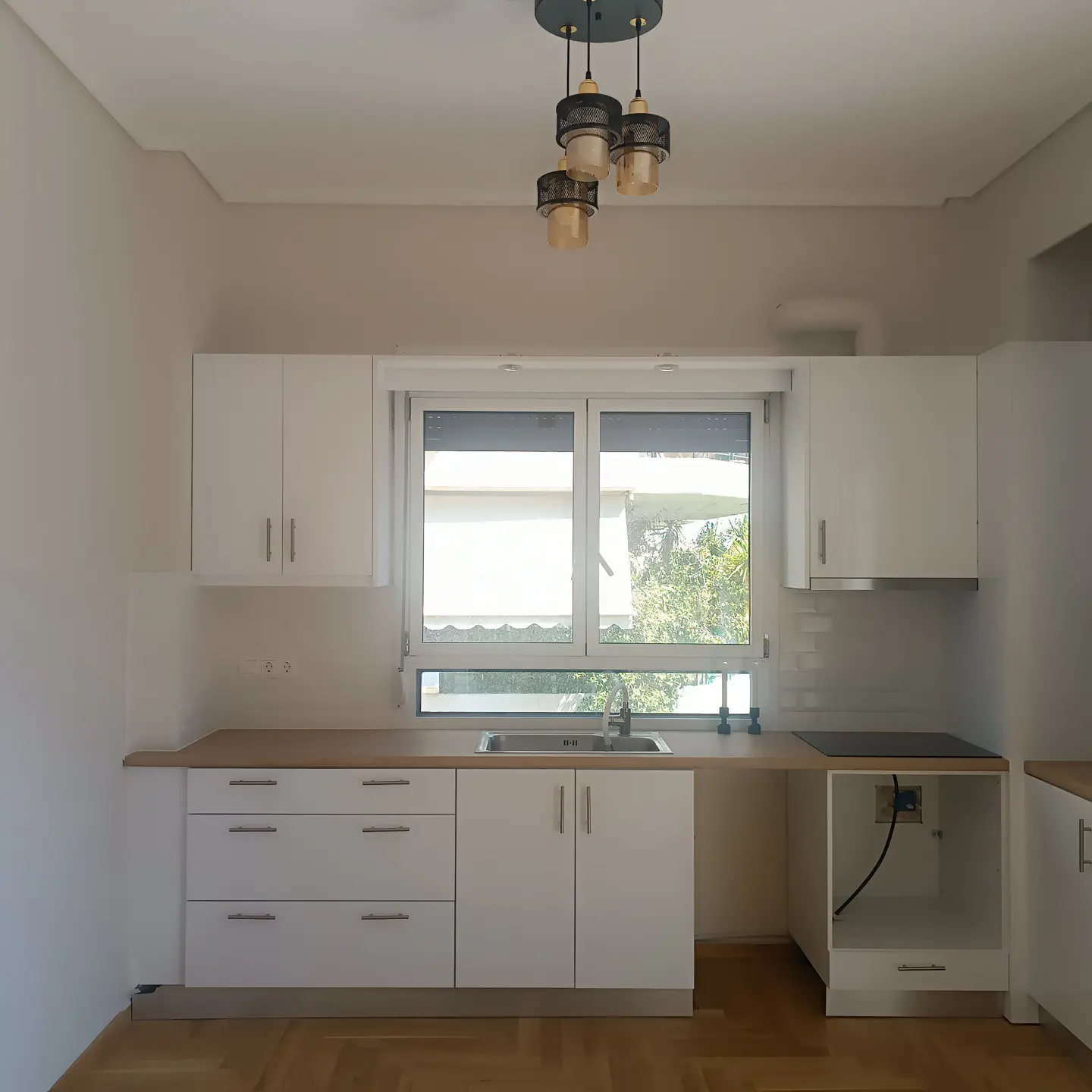 Bright kitchen with white cabinets, wood countertops, and herringbone floors. A window overlooks greenery. A modern chandelier hangs above.