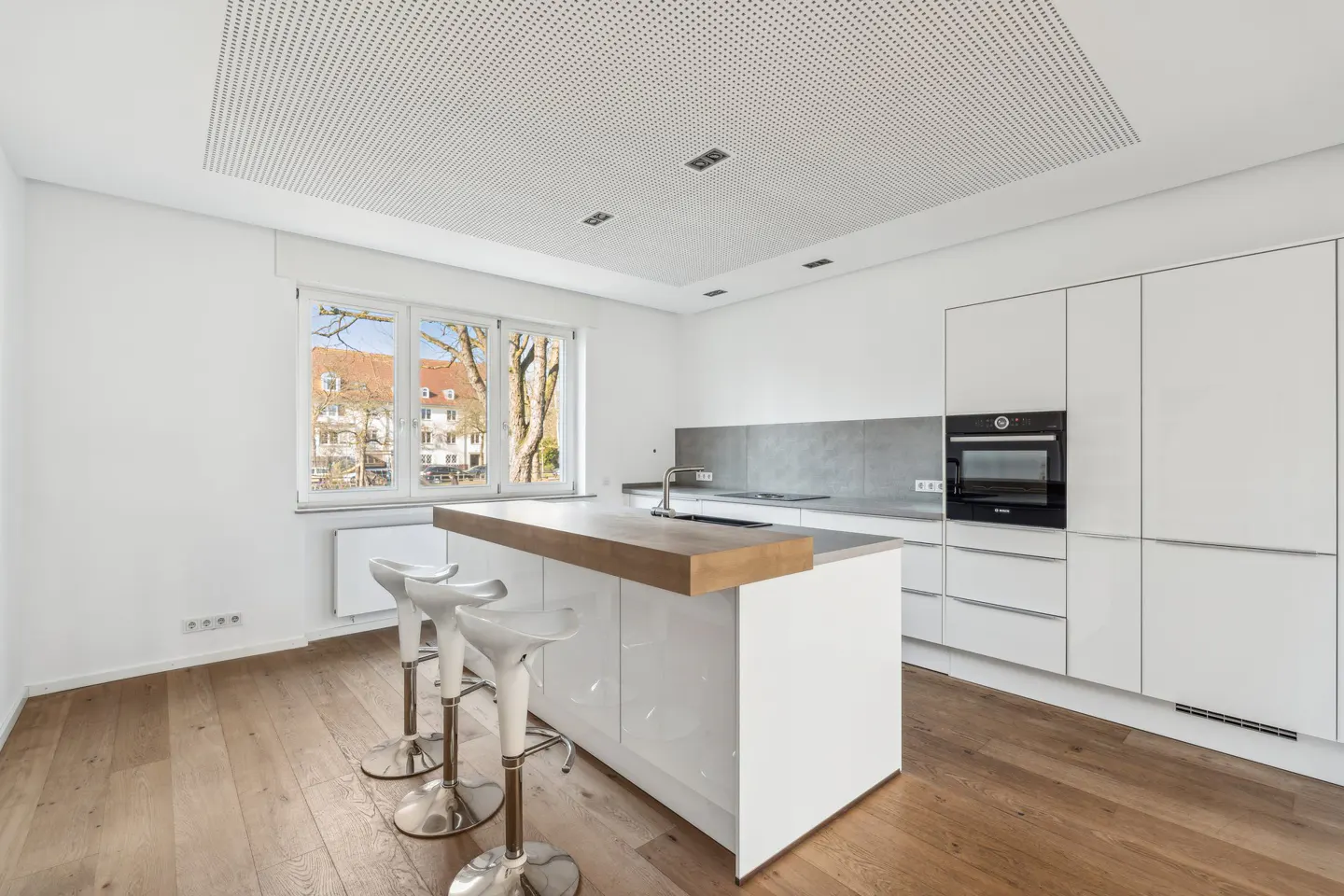 Bright, modern kitchen with white cabinets, wood floors, and a center island with three white bar stools. A window overlooks a building.