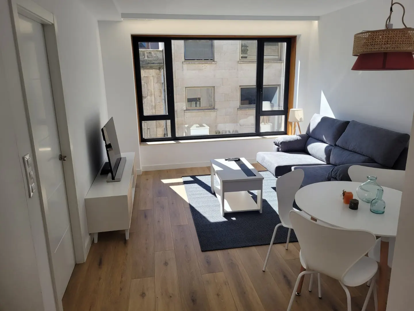 Bright living room with wood floors, a gray sofa, white table and chairs, and a large window with a city view.