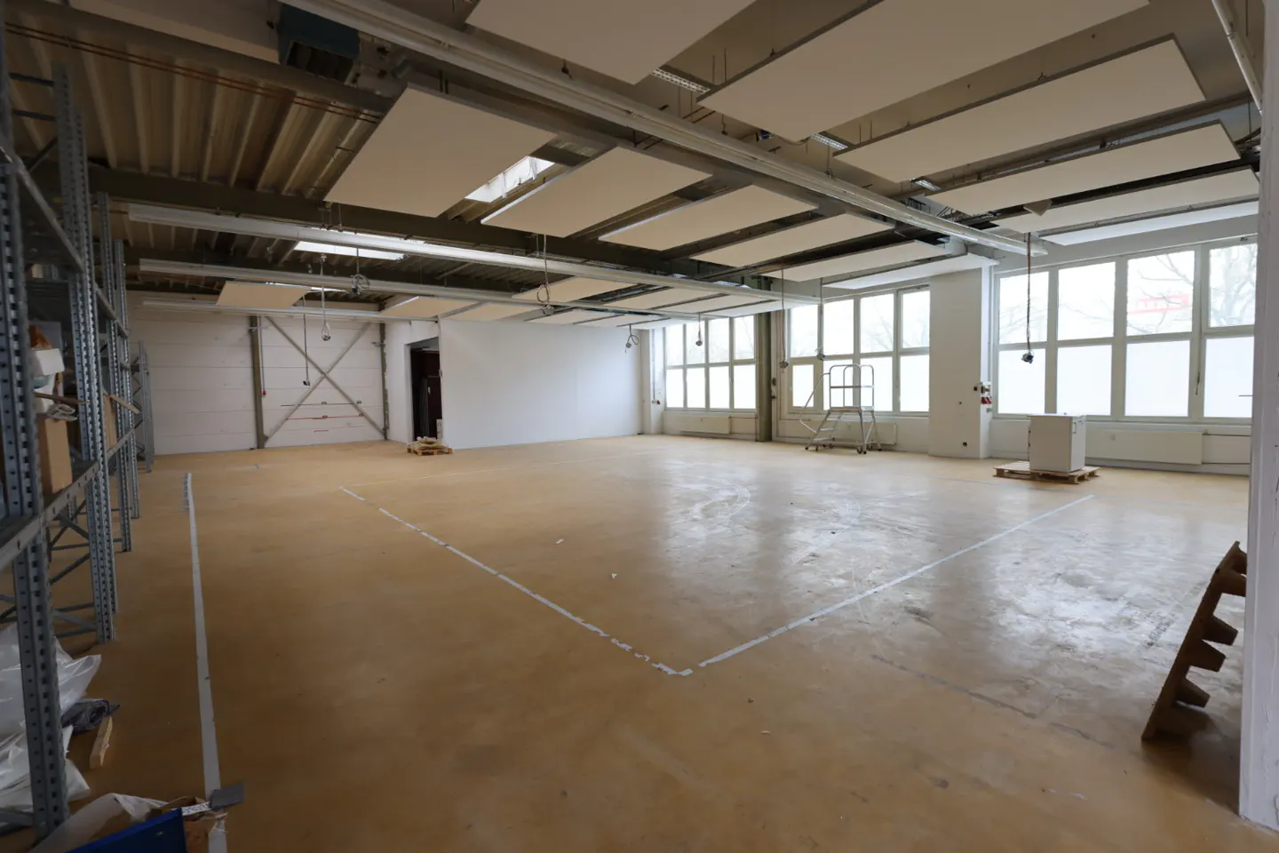 Large, empty commercial space with beige floor, white walls, and suspended ceiling panels. Windows line the back wall.
