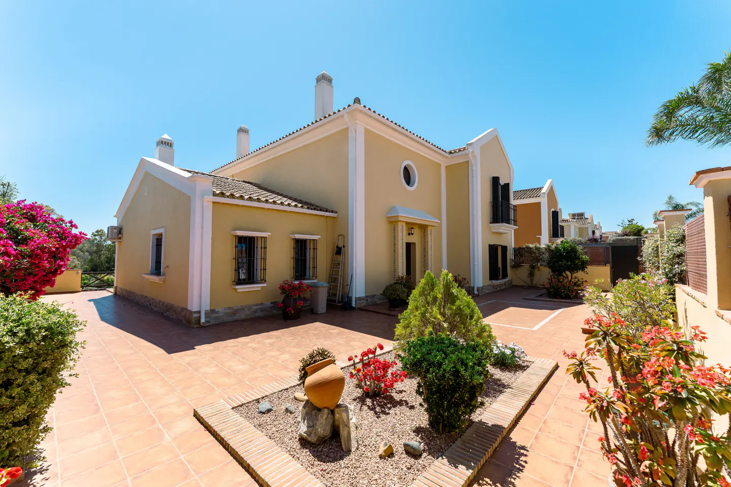 Exterior view of a two-story, tan house with white trim, a tiled roof, and a brick-paved courtyard with flower beds.
