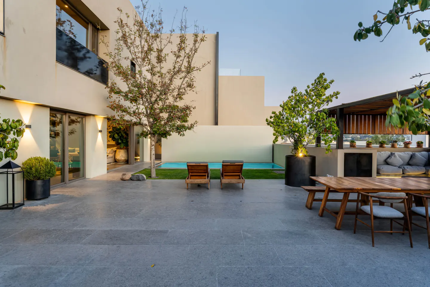 Modern home exterior with a pool, lounge chairs, and outdoor dining area on a gray stone patio. Trees and plants add greenery.