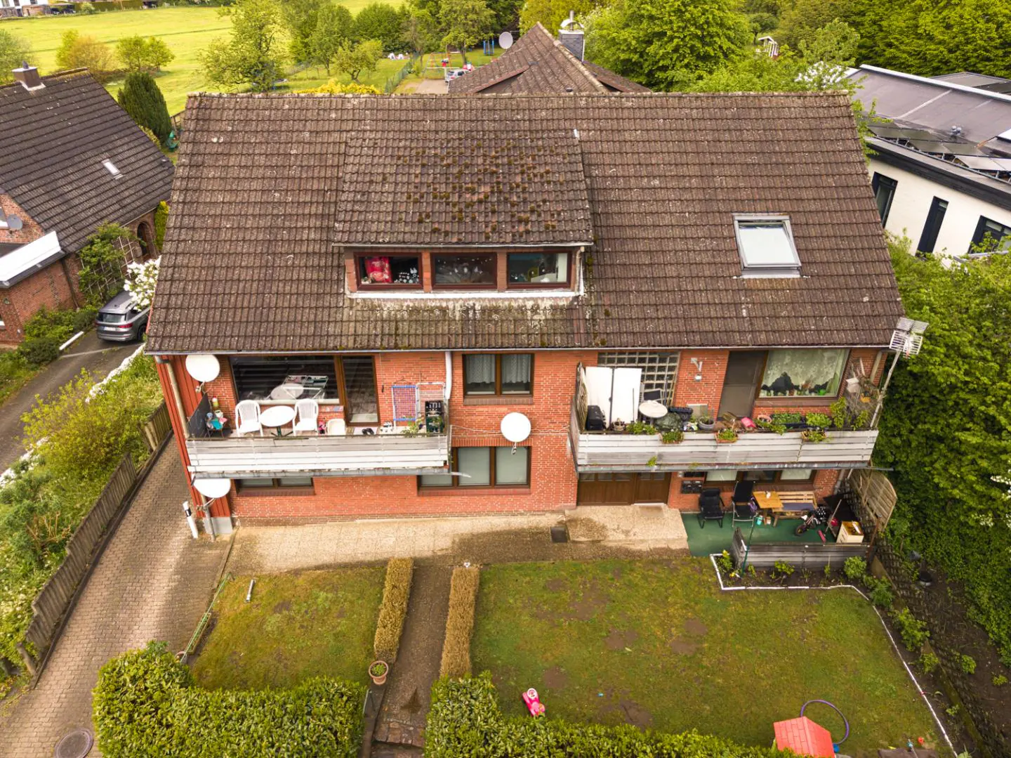 Aerial view of a two-story red brick house with a brown tiled roof, two balconies, and a green backyard.
