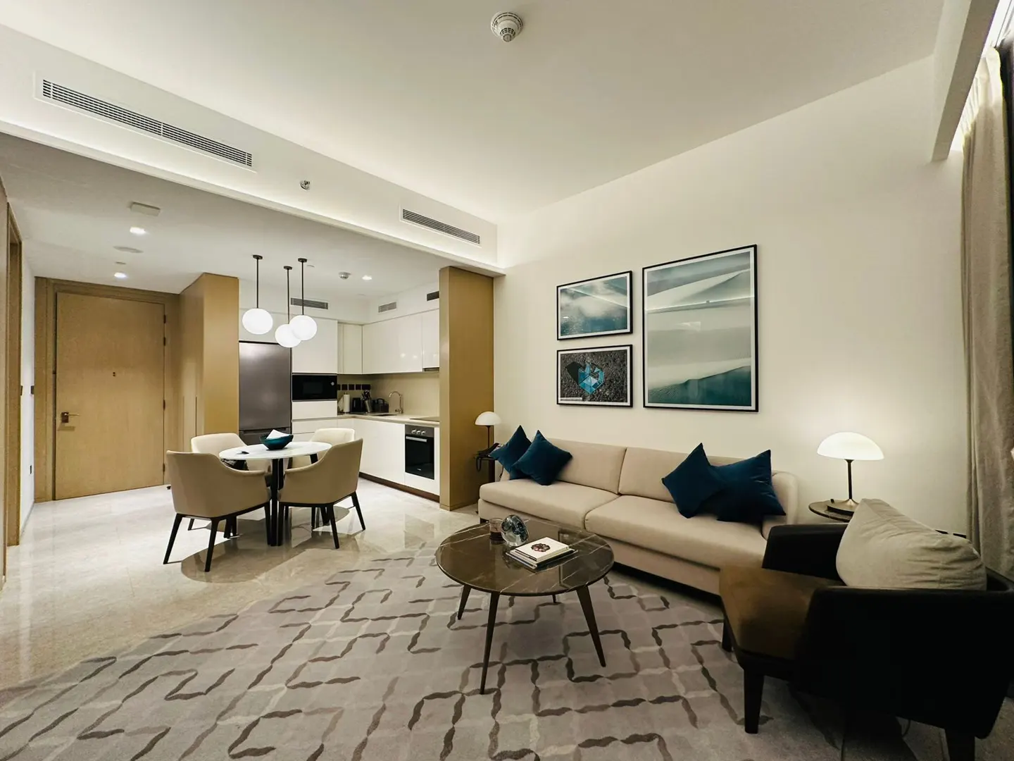 A modern apartment interior with a beige sofa, blue pillows, and a geometric patterned rug. A dining area and kitchen are visible in the background.