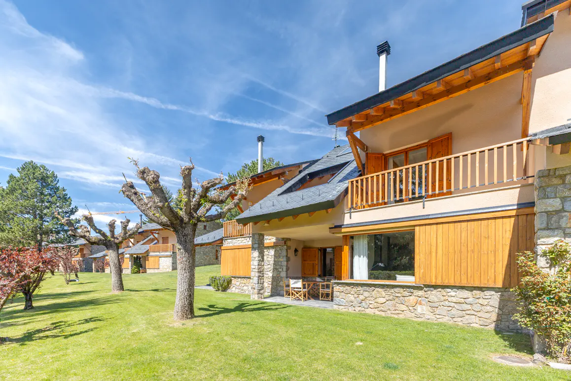 Exterior view of a tan house with wood and stone accents, a balcony, and a green lawn under a blue sky.