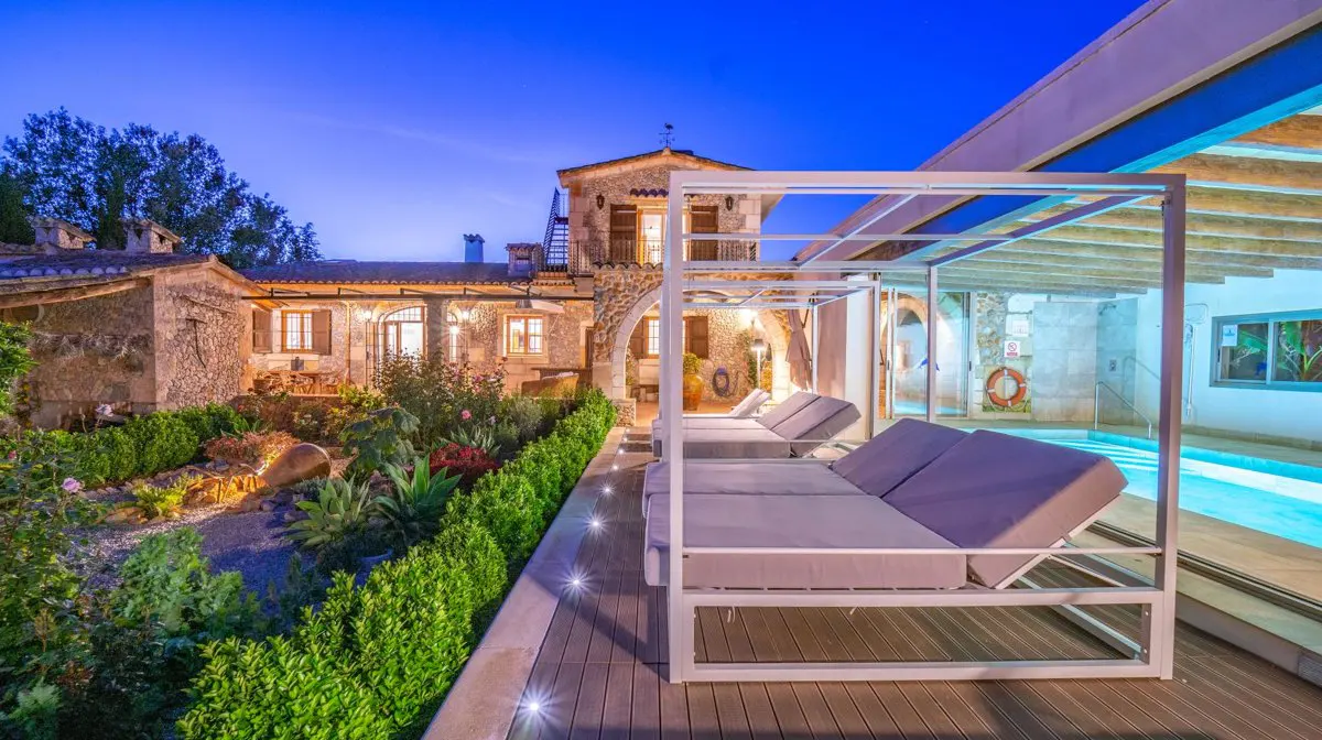Outdoor view of a stone building with a pool and lounge chairs at dusk. The building has lit windows and a garden.