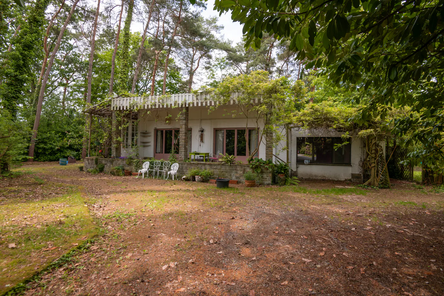 A white house with pink trim is surrounded by trees and greenery. A porch with white chairs sits in front.