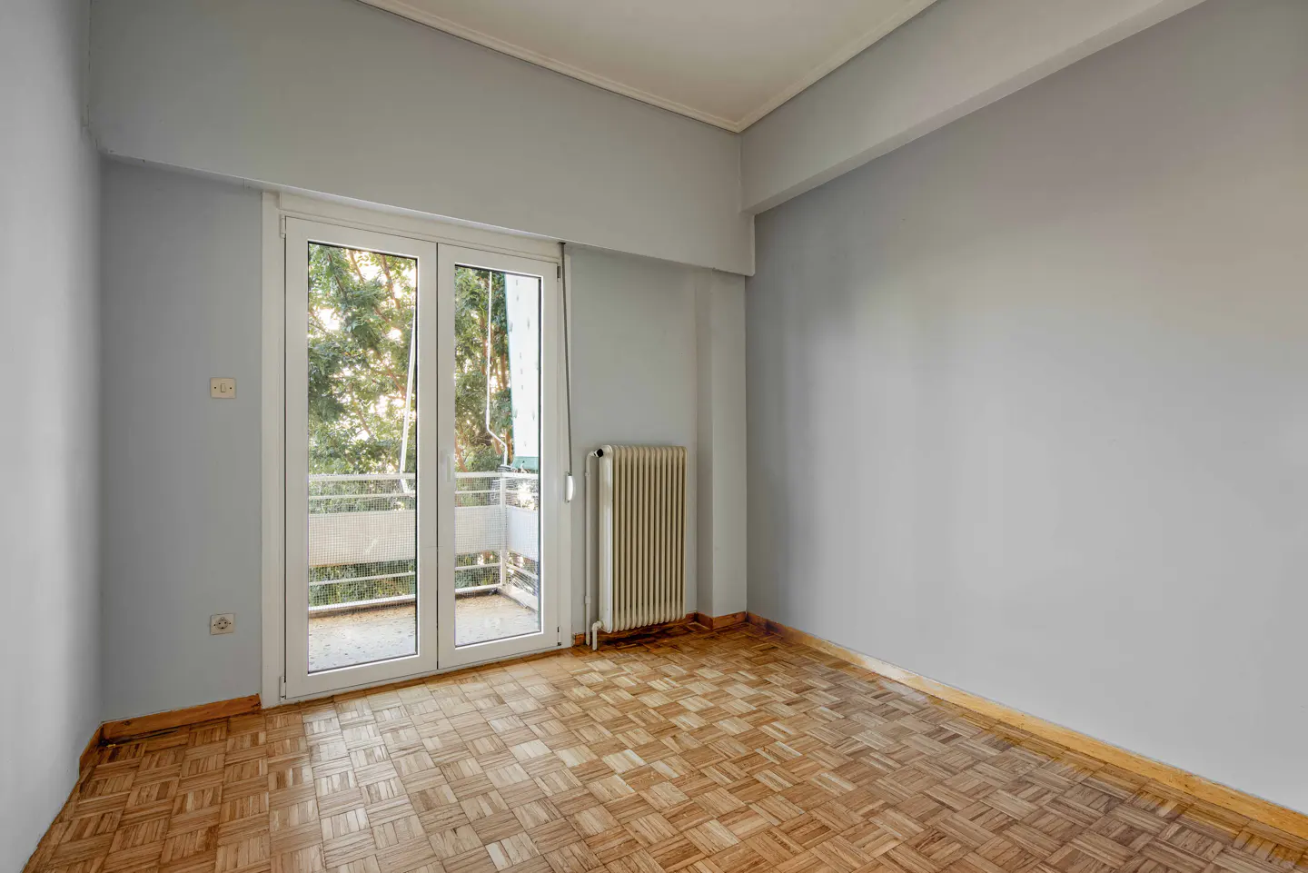 Empty room with parquet floor, light gray walls, and white-framed glass doors leading to a balcony with trees visible.