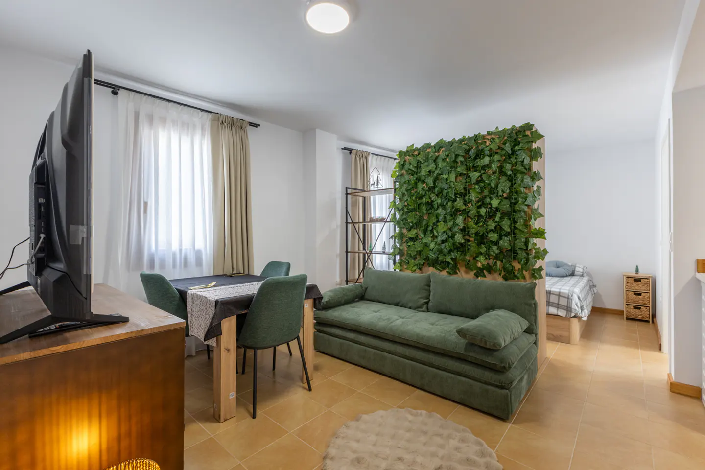 Living room with green sofa, dining table, and TV. A wall of ivy separates the living area from a bedroom with a bed and nightstand.
