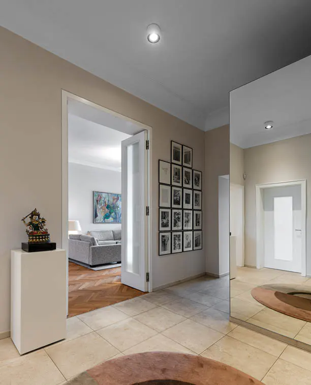 Interior view of a home's entrance hall with beige walls, tile flooring, and a large mirror. A doorway leads to a living room with a gray sofa and artwork. Black and white photos adorn the wall.