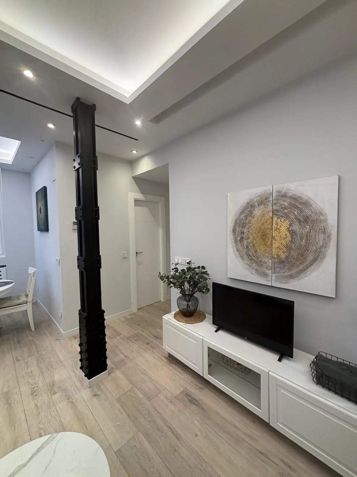 Modern living room with light wood floors, gray walls, and white trim. A black support beam stands near a dining area. A TV sits on a white console under abstract art.
