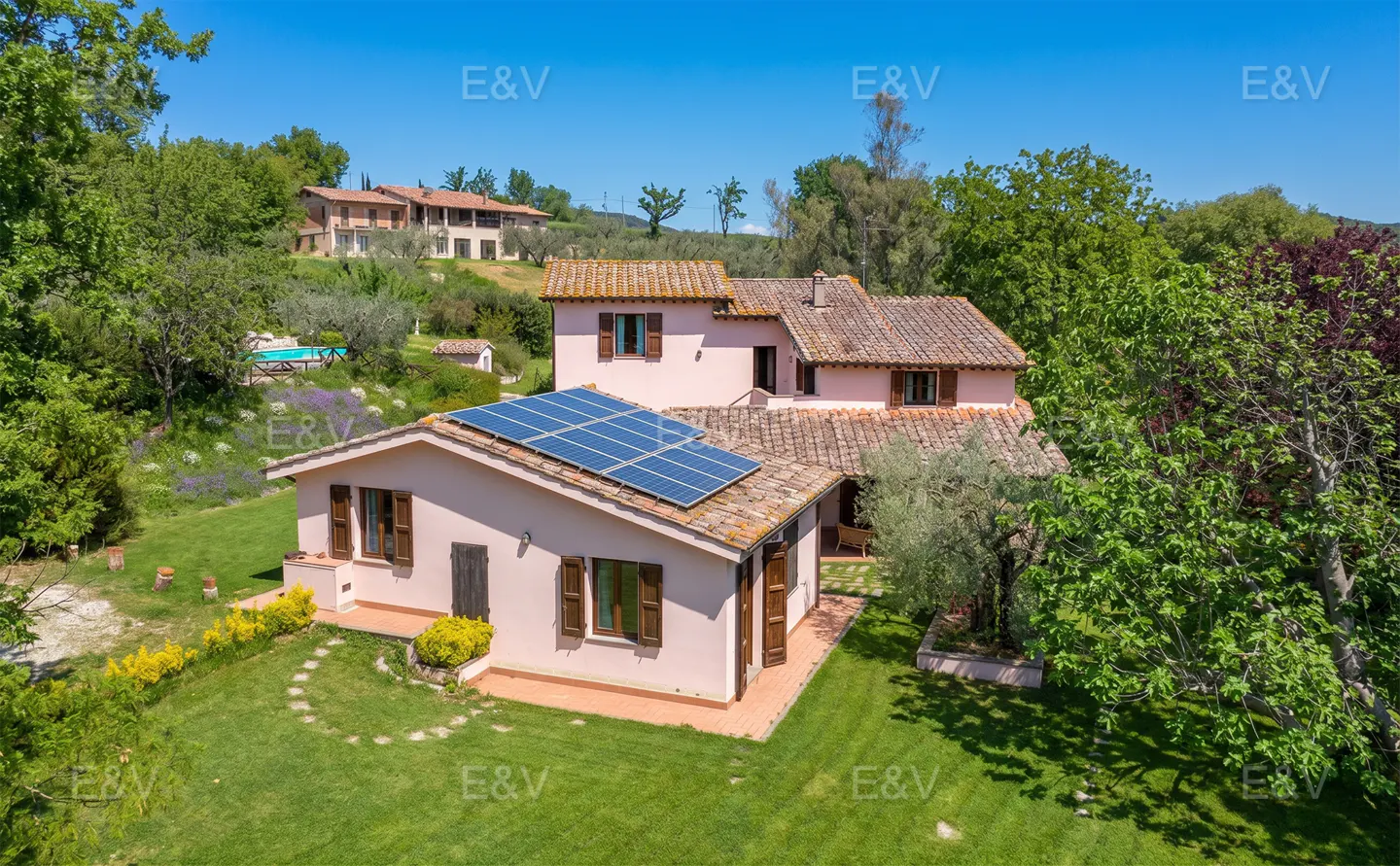 Aerial view of a pink two-story house with brown shutters and a solar panel roof, surrounded by green trees and grass.