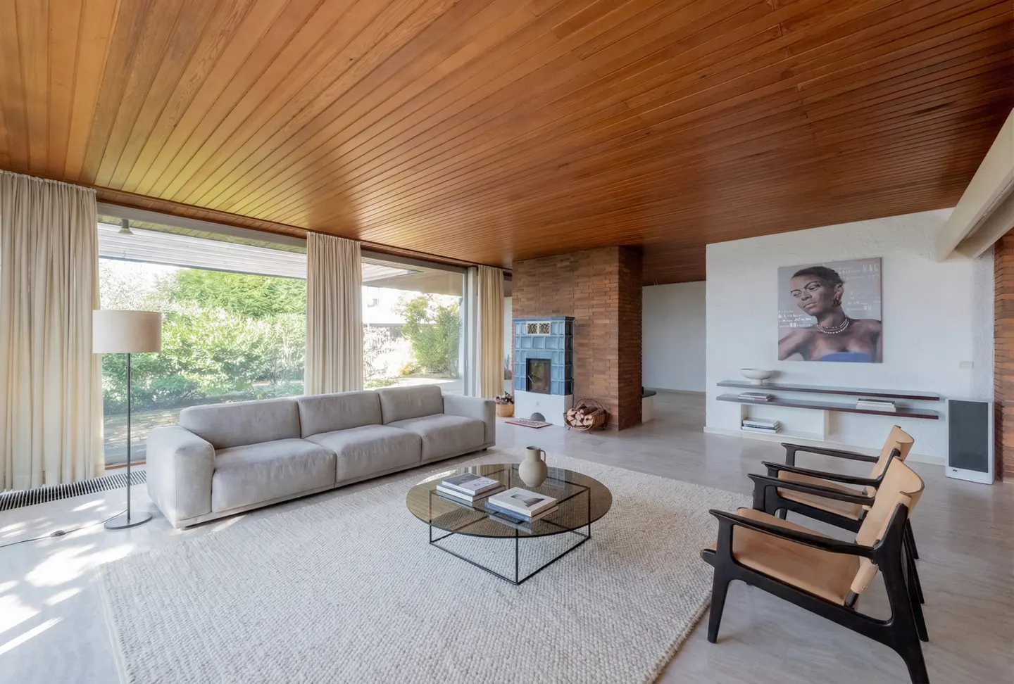Living room with wood ceiling, large window, and gray sofa. A glass table sits on a white rug. Two chairs face a painting on the wall.