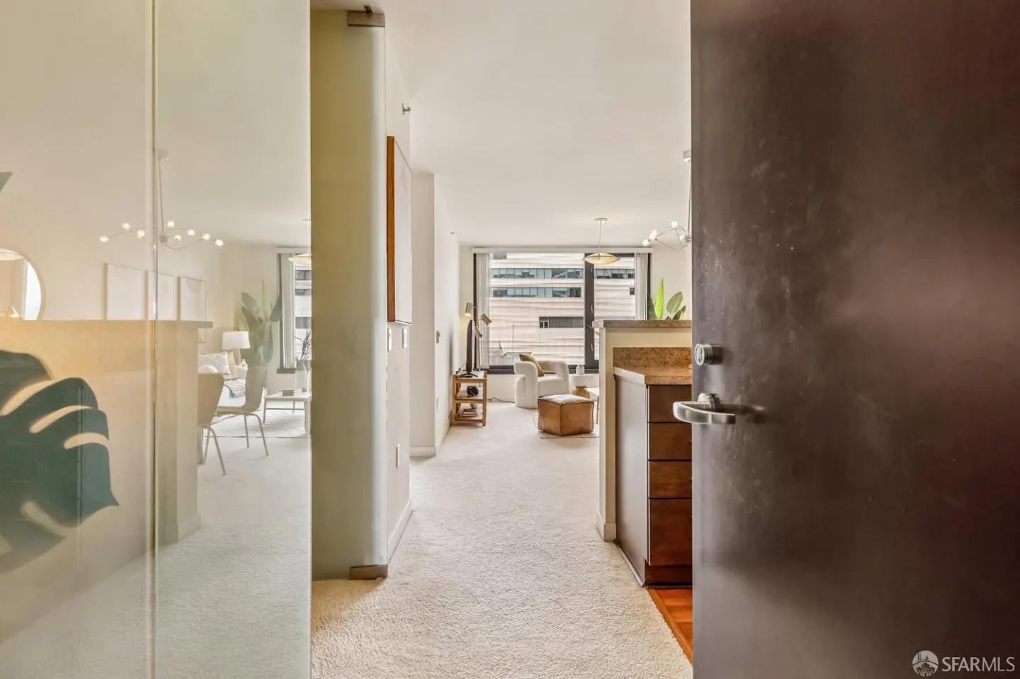 View from a dark doorway into a bright, carpeted apartment living area with a glass wall and city view through windows.