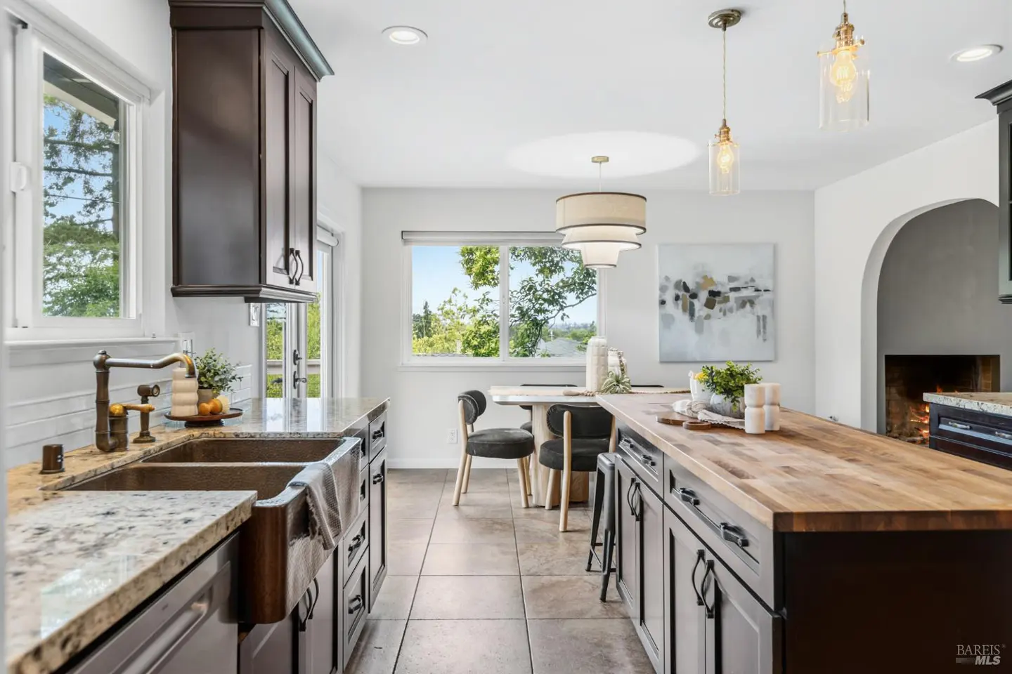 Modern kitchen with dark cabinets, granite countertops, a wood-topped island, and a dining area with a view of trees.