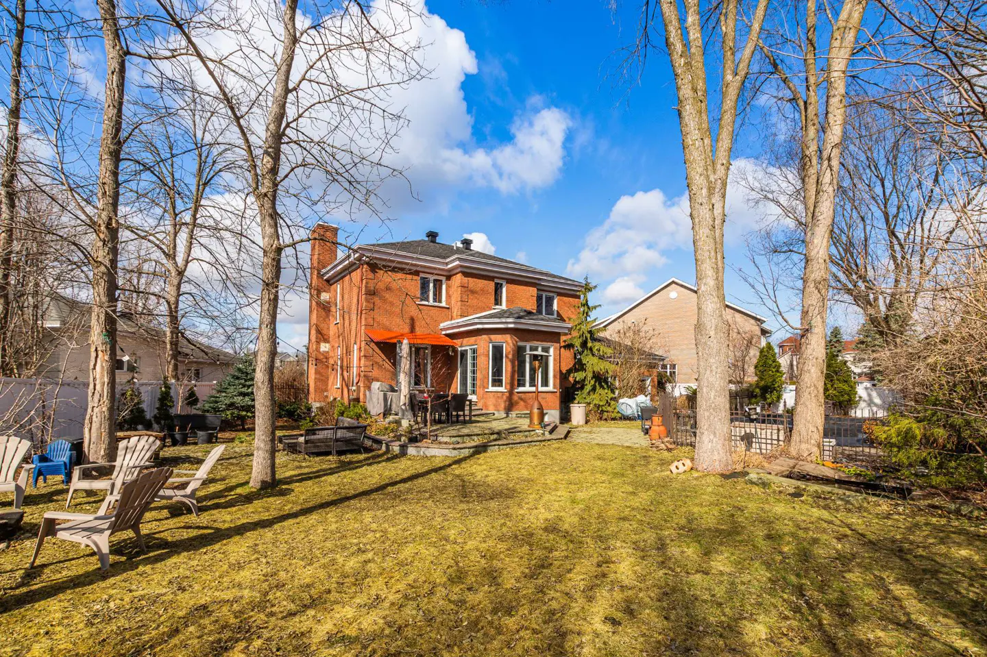 Backyard view of a two-story red brick house with a patio, lawn, and trees under a blue sky with clouds.
