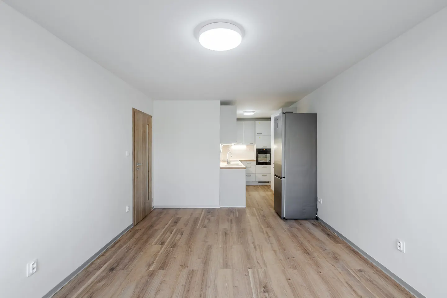 Bright, empty room with wood floors, white walls, and a kitchen area with a stainless steel refrigerator.
