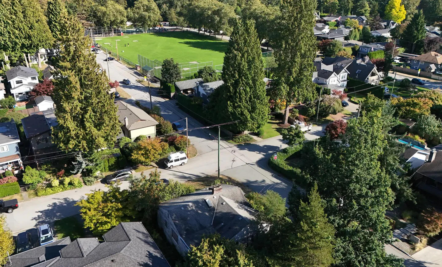 Aerial view of a residential neighborhood with green trees, houses, and a green sports field in the background.