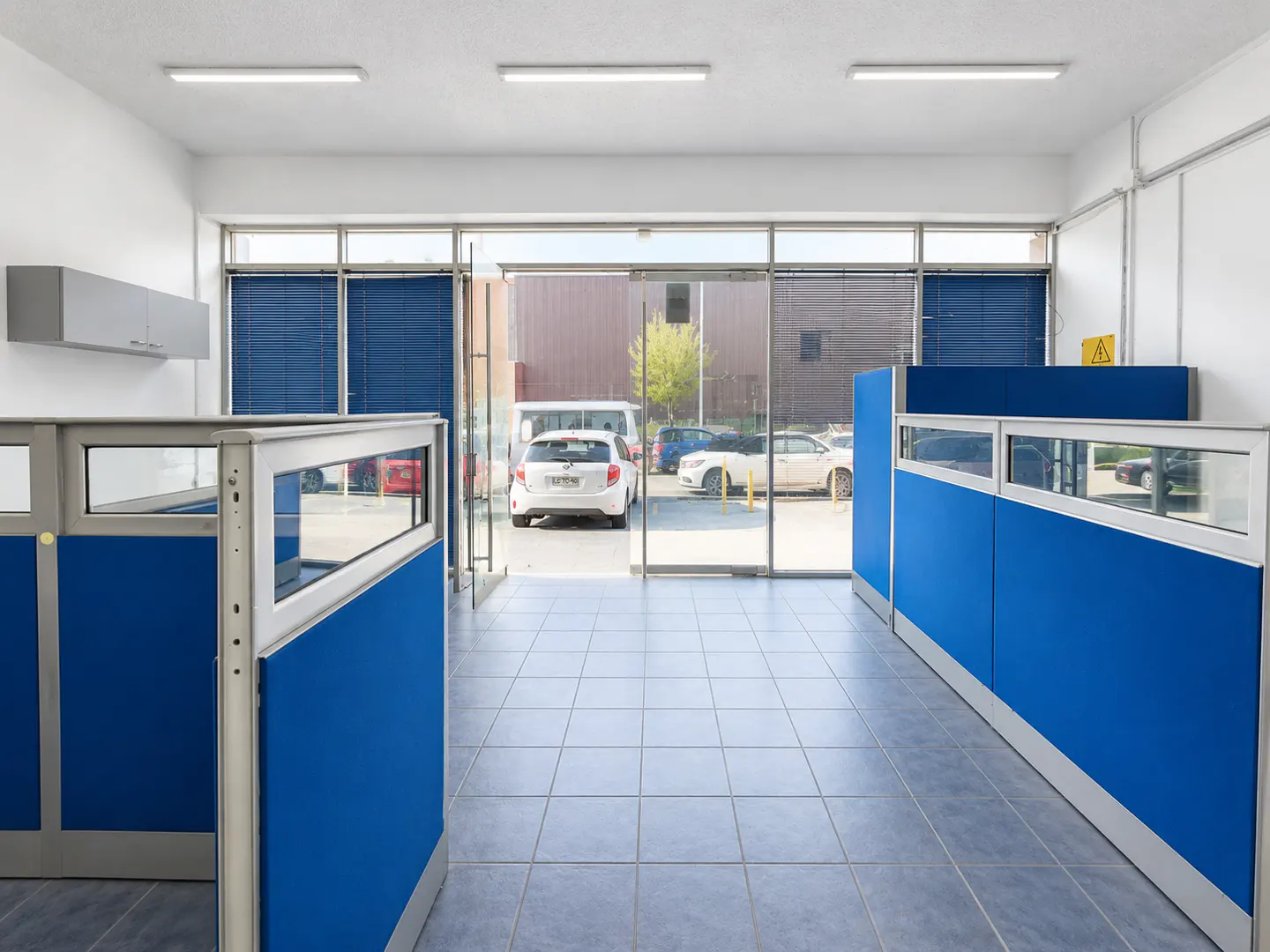 Office interior with blue cubicle partitions, tiled floors, and a large glass wall looking out to a parking lot.