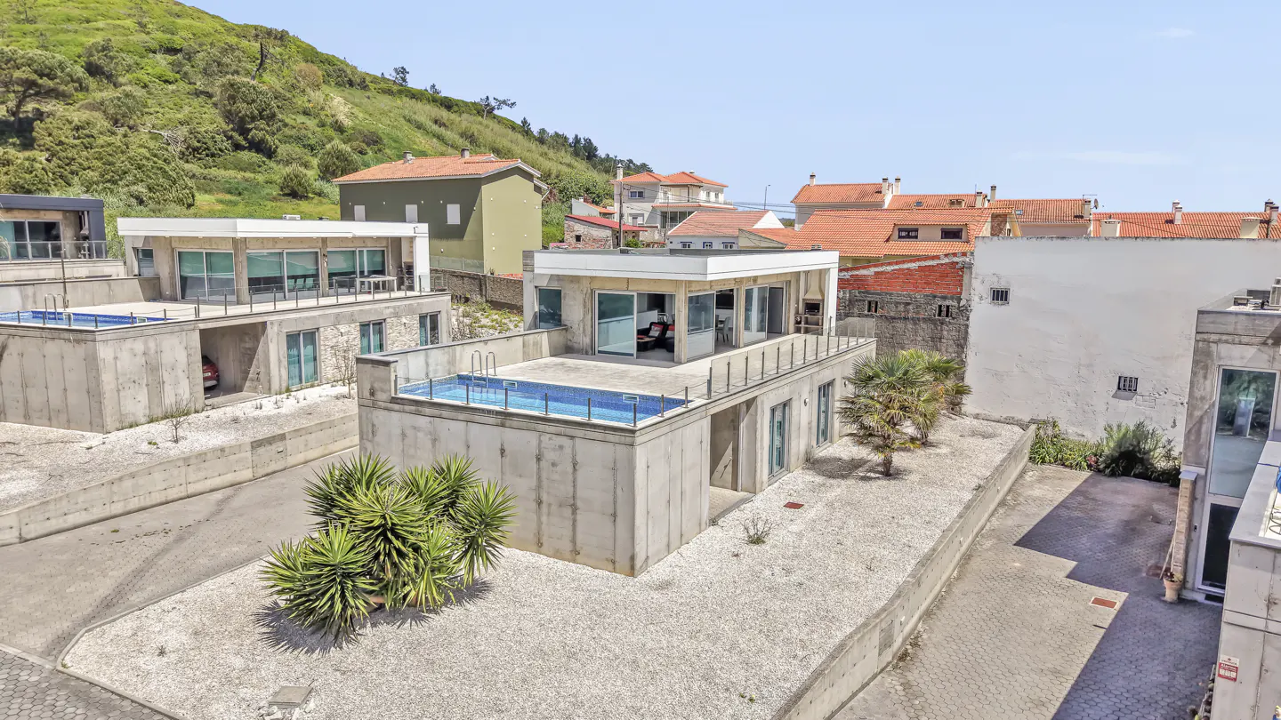 Aerial view of modern concrete homes with rooftop pools, surrounded by greenery and red-roofed buildings under a blue sky.