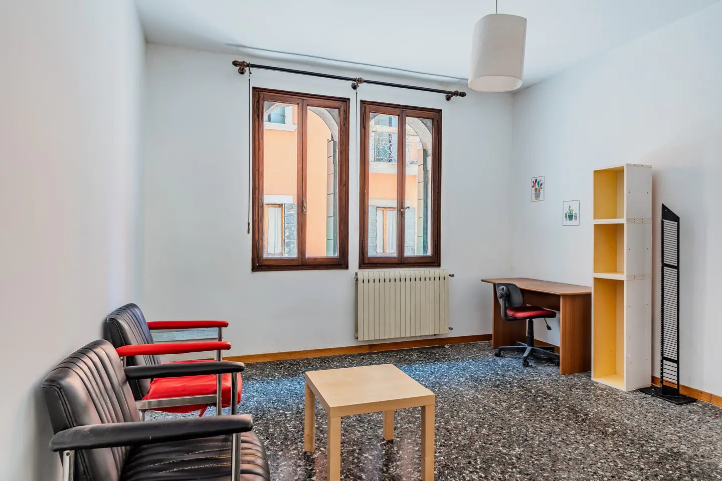 Bright room with two black and red chairs, a light wood table, and a desk with a red chair. Two windows with brown frames look out onto a building.