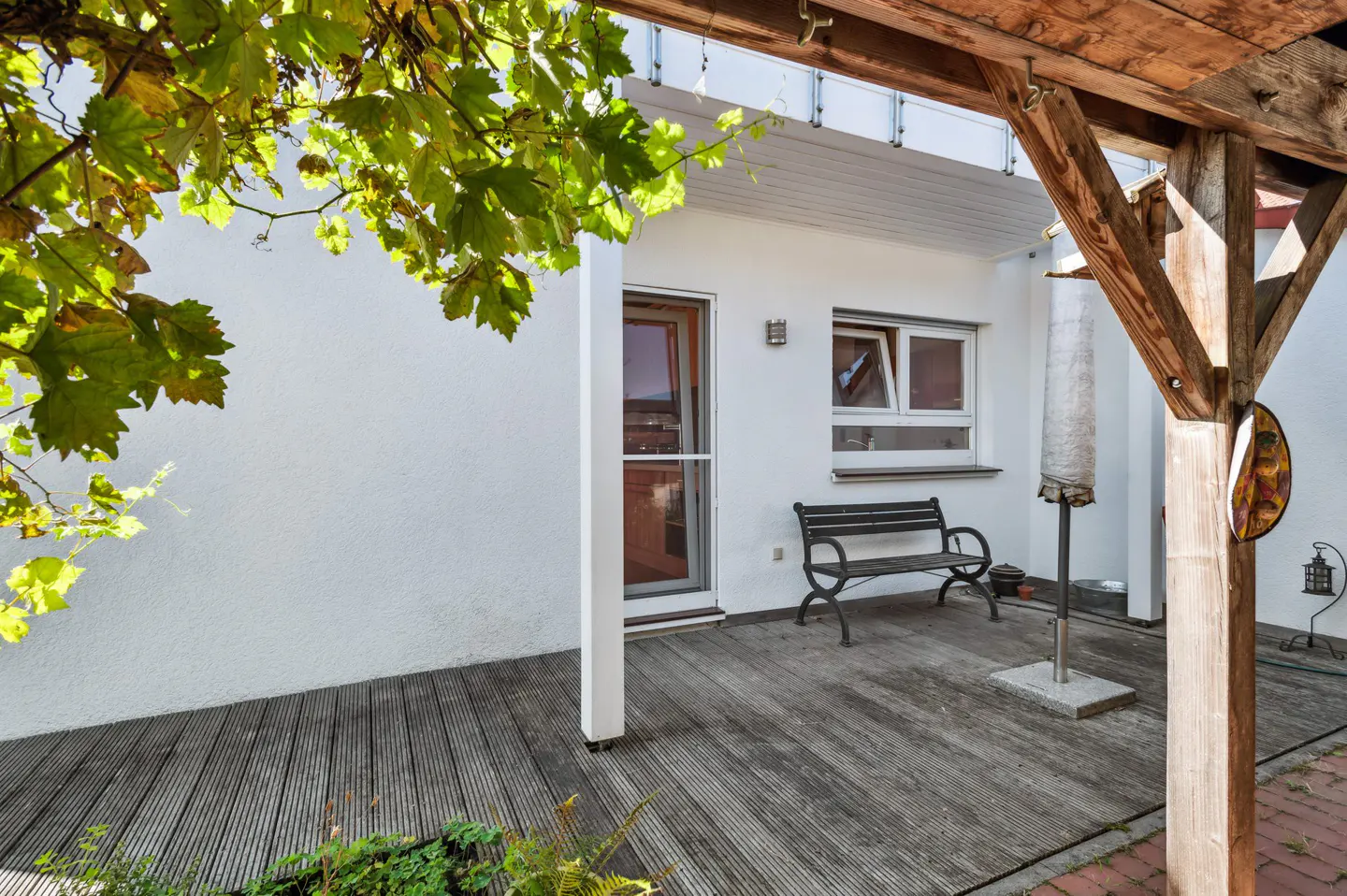Outdoor patio with wooden deck, white walls, and a wooden pergola. A black bench sits near a window, and green leaves hang overhead.