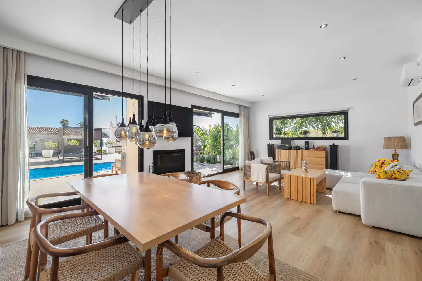 Bright, modern living room with a wood dining table, chairs, and pendant lights. Sliding glass doors open to a pool area. White sofa and wood furniture.