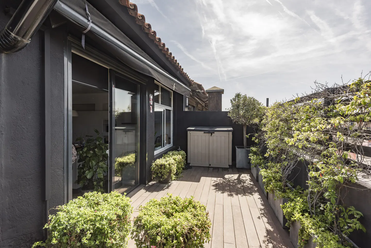 A rooftop patio with a wooden deck, green plants in pots, and a storage box against a dark gray building.