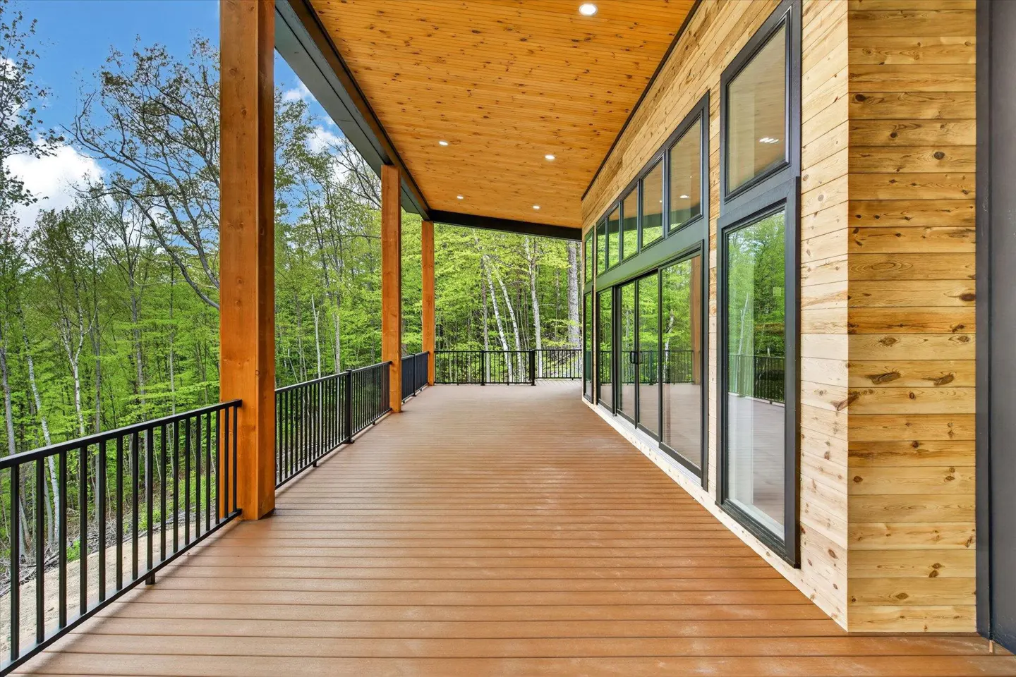 Wide porch with wood floors, black railings, and wood pillars overlooking a green forest. Large windows line the wood-paneled wall.
