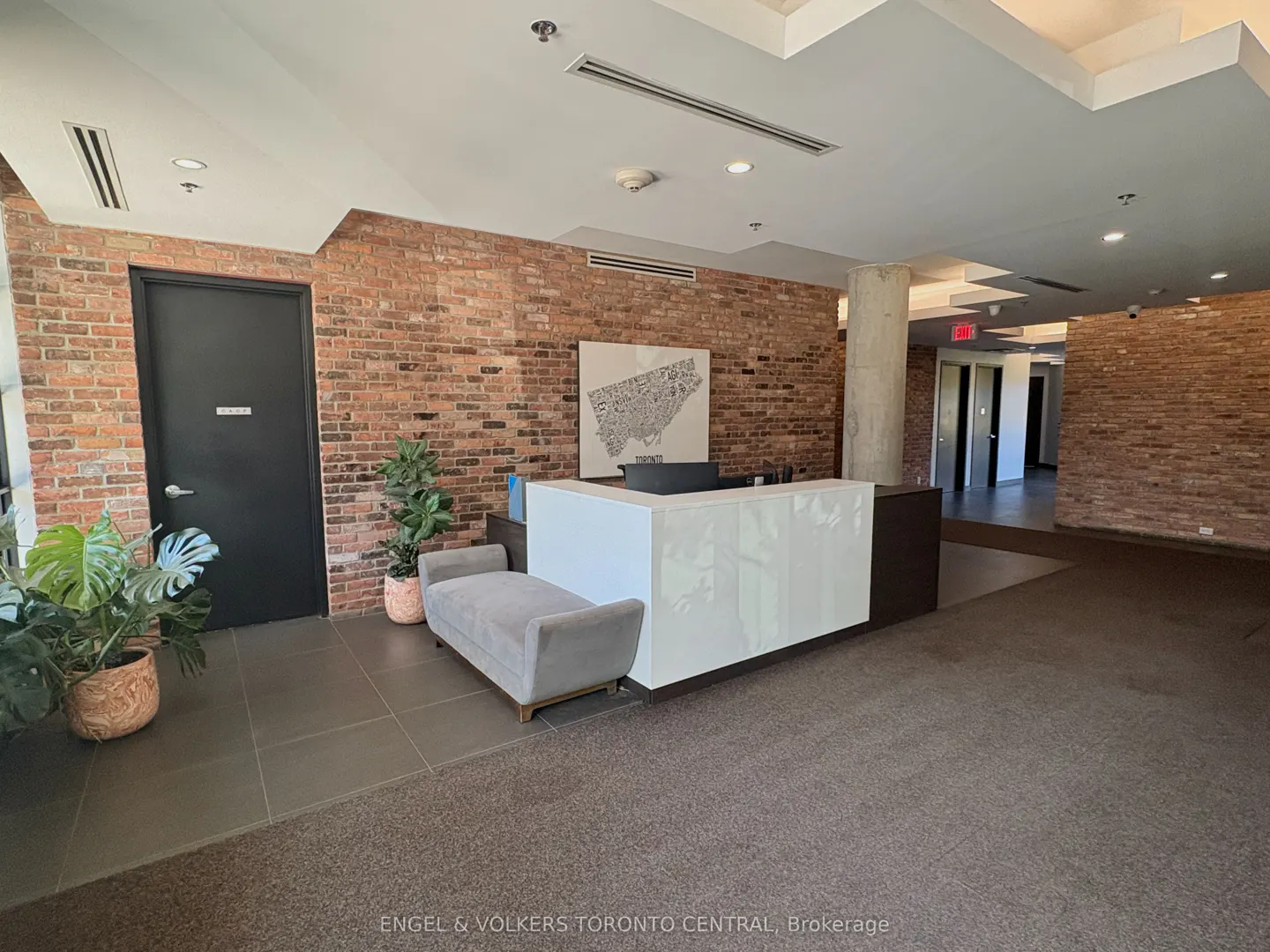 Condo lobby with a white reception desk, grey bench, and brick walls. A Toronto map hangs above the desk. Plants add a touch of greenery.