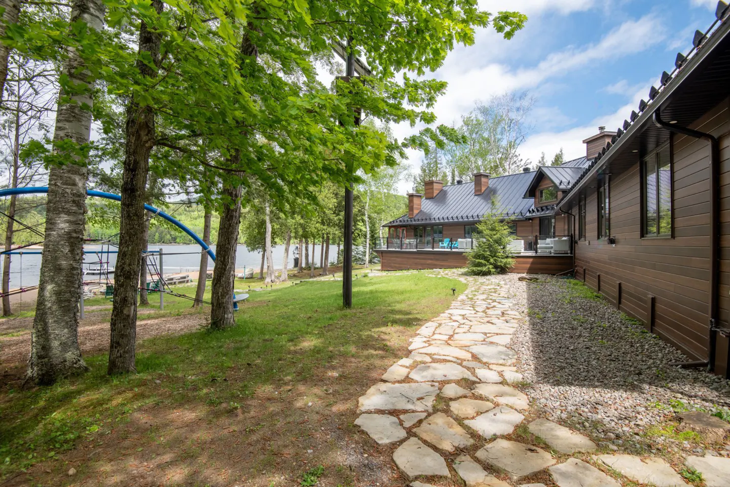 Stone path leads to a brown house with a black roof, near a lake with a playground and green trees.
