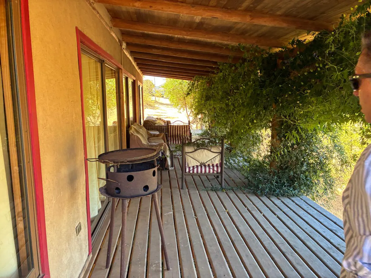 Covered porch with wooden floors, grill, and seating. A man in sunglasses stands to the right. Green foliage and trees are visible in the background.