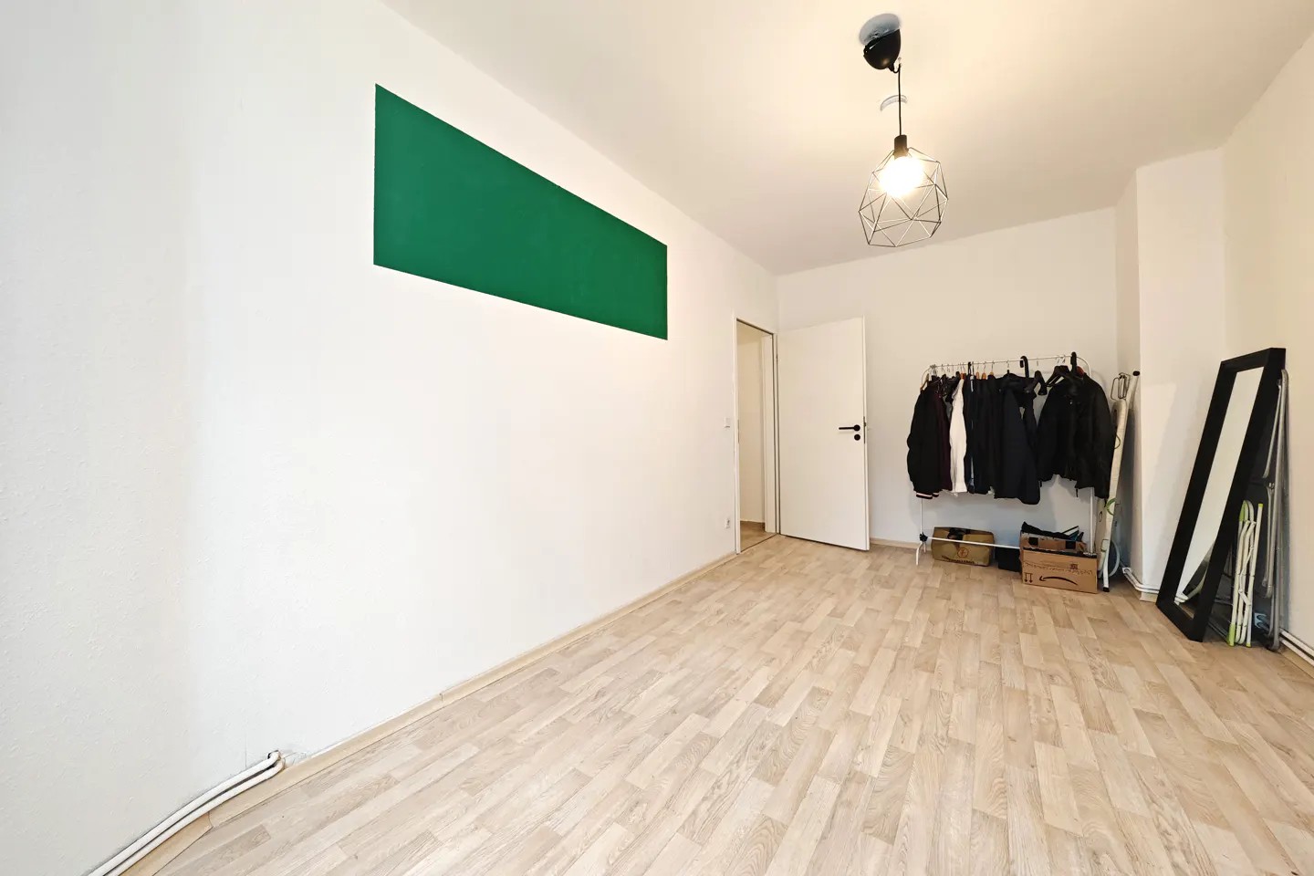 Bright room with wood floors, white walls, and a green rectangle. Clothes hang on a rack near a mirror and an open white door.