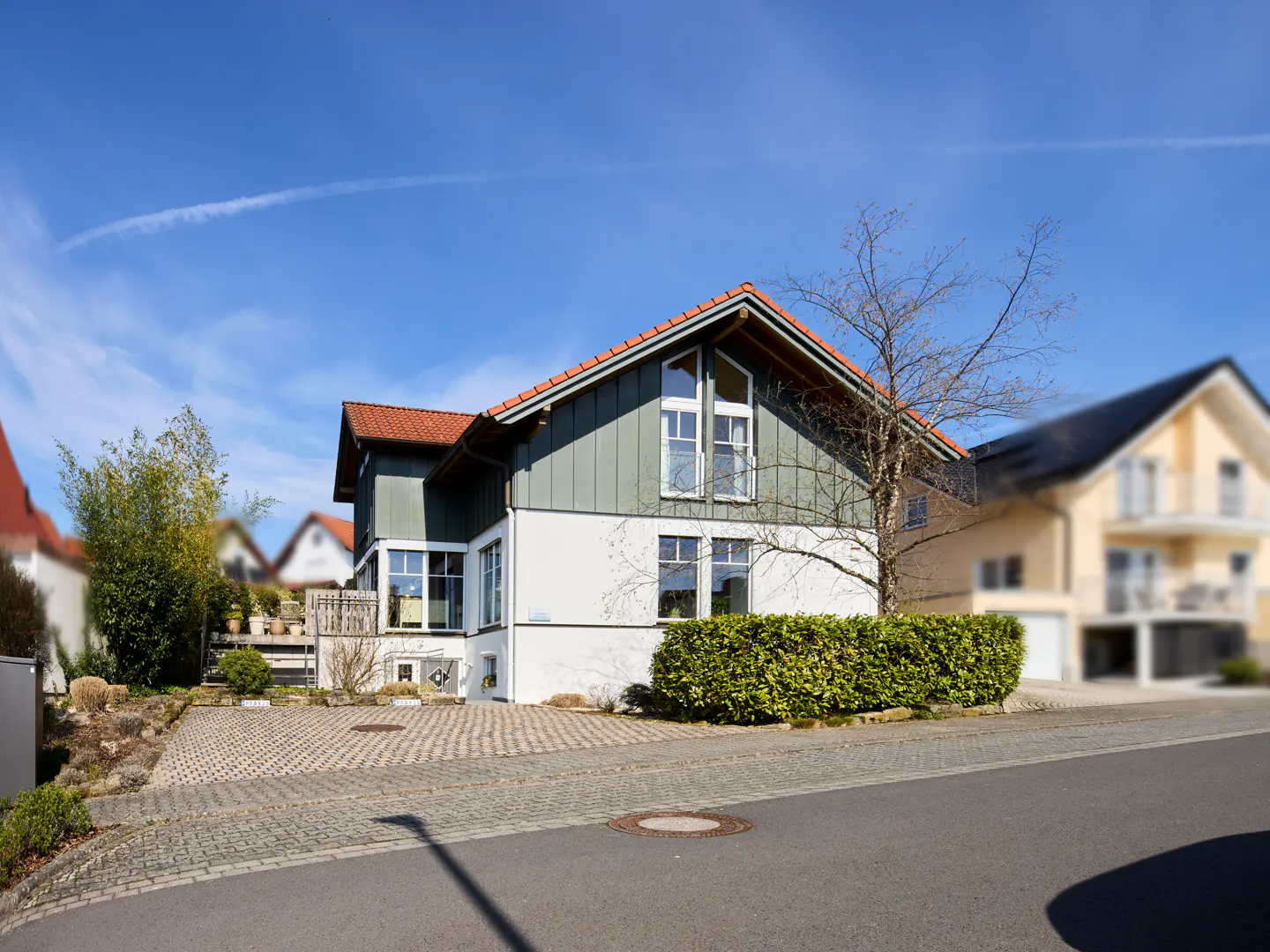Two-story house with green siding, white stucco, and a red tile roof on a sunny day. A green hedge lines the front of the house.