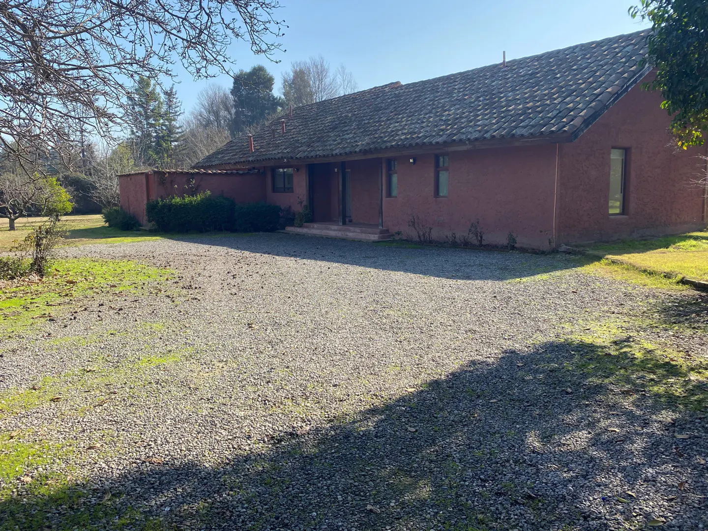 A single-story, terracotta-colored house with a tiled roof sits behind a large gravel driveway on a sunny day.