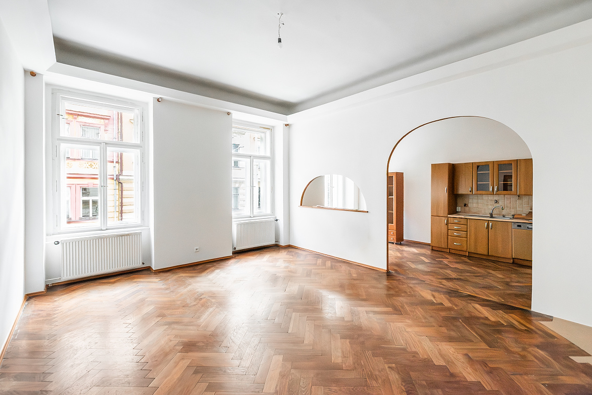 Bright living room with herringbone wood floors, white walls, large windows, and an arched doorway leading to a kitchen. [AI-generated text]
