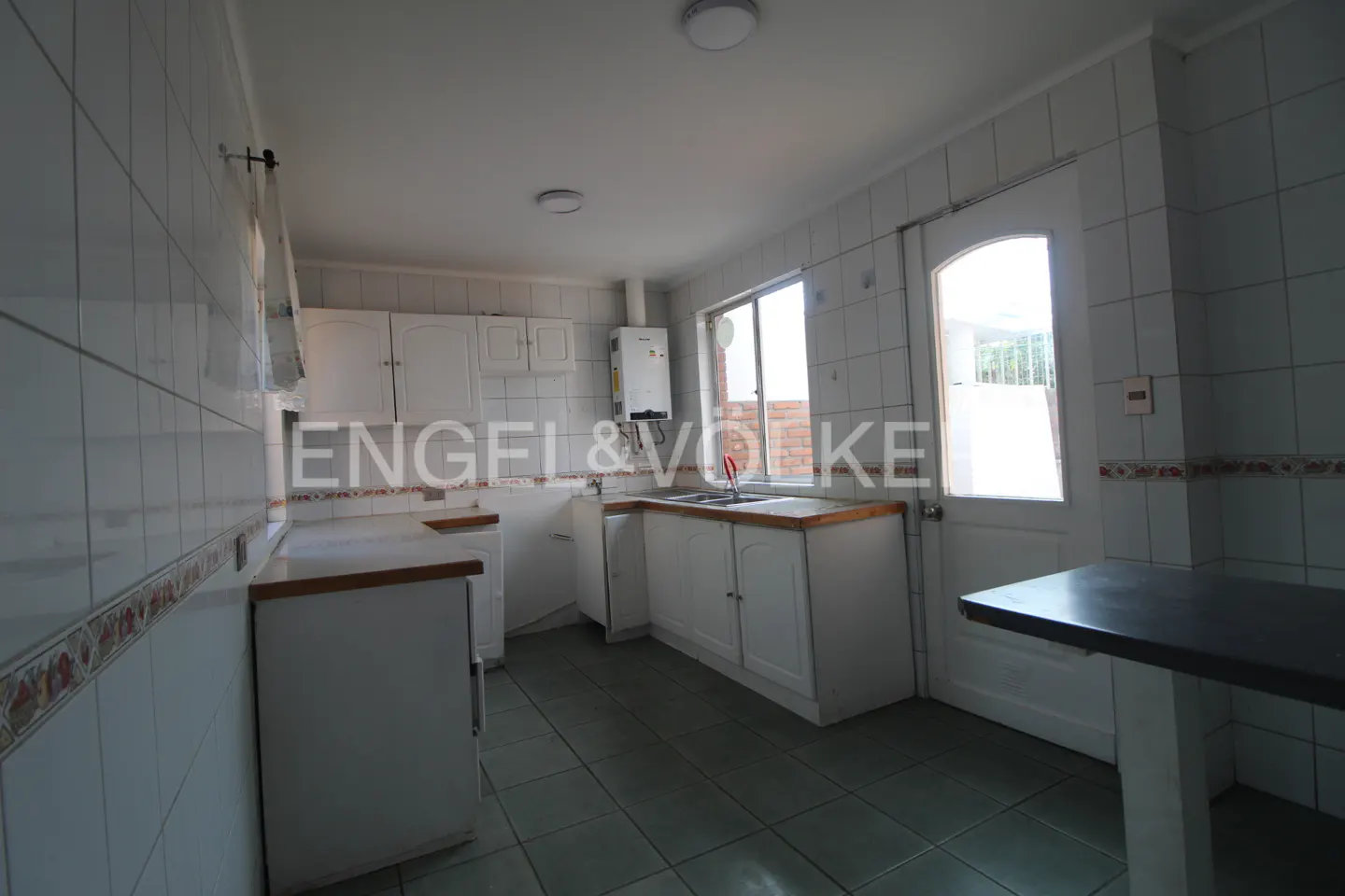 A bright kitchen with white cabinets, tiled walls, and a window. A dark table stands on the right.