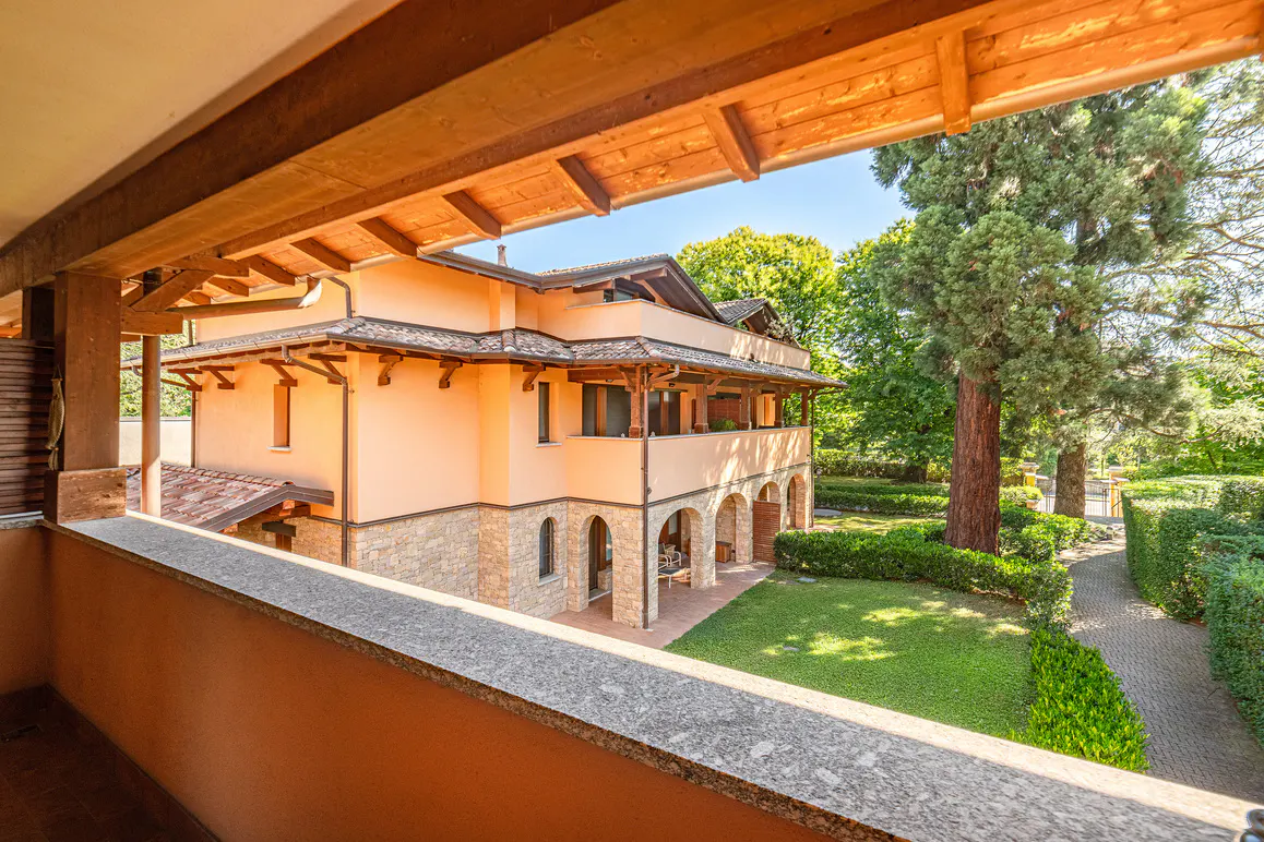 View from a balcony overlooking a peach-colored villa with stone accents, a green lawn, and tall trees on a sunny day.
