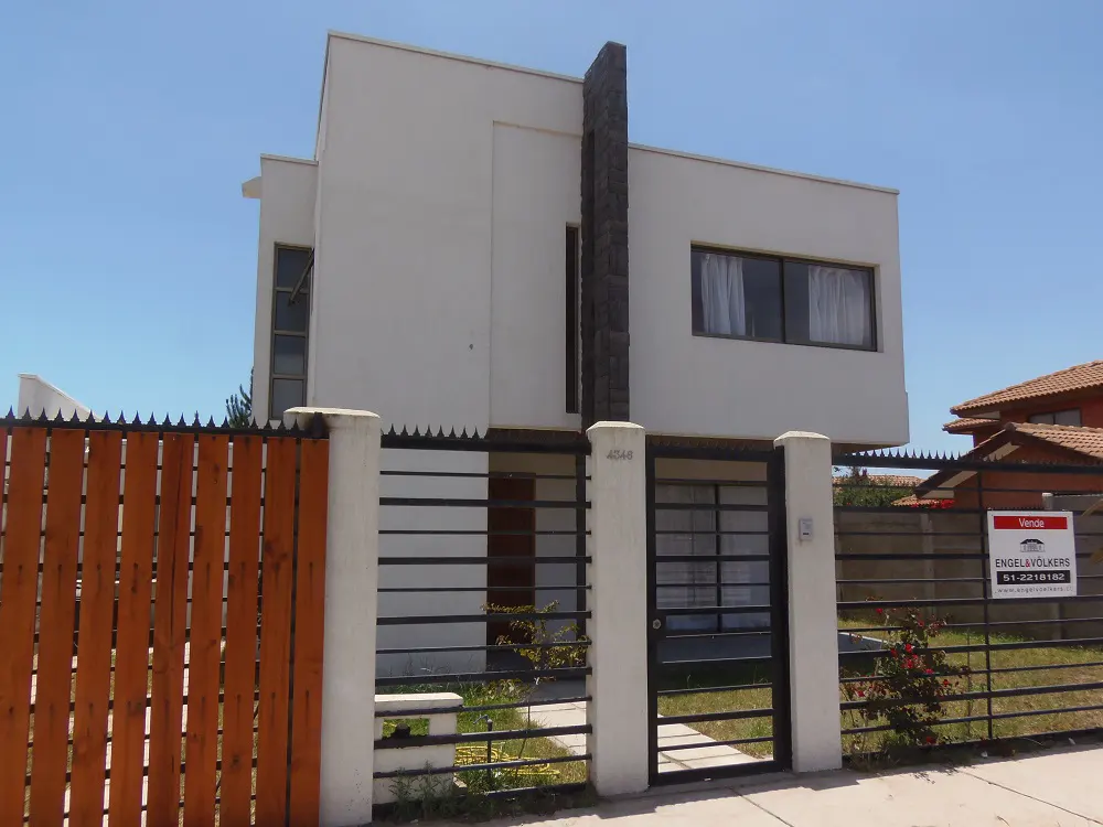 Two-story modern white house with a dark stone accent, behind a black metal and wood fence. A "For Sale" sign is visible.
