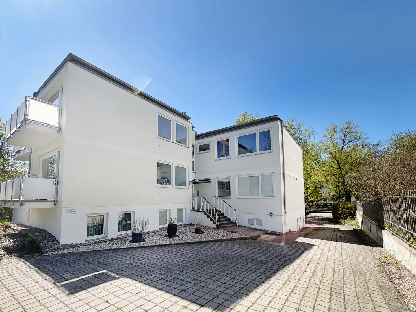 Exterior view of a two-story white apartment building with balconies and a paved driveway under a clear blue sky.