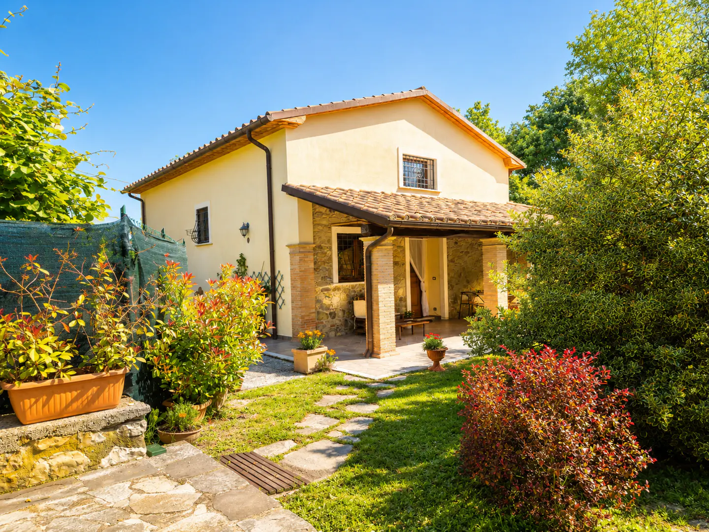 Two-story yellow house with a tiled roof and covered porch, surrounded by lush green landscaping and a blue sky.