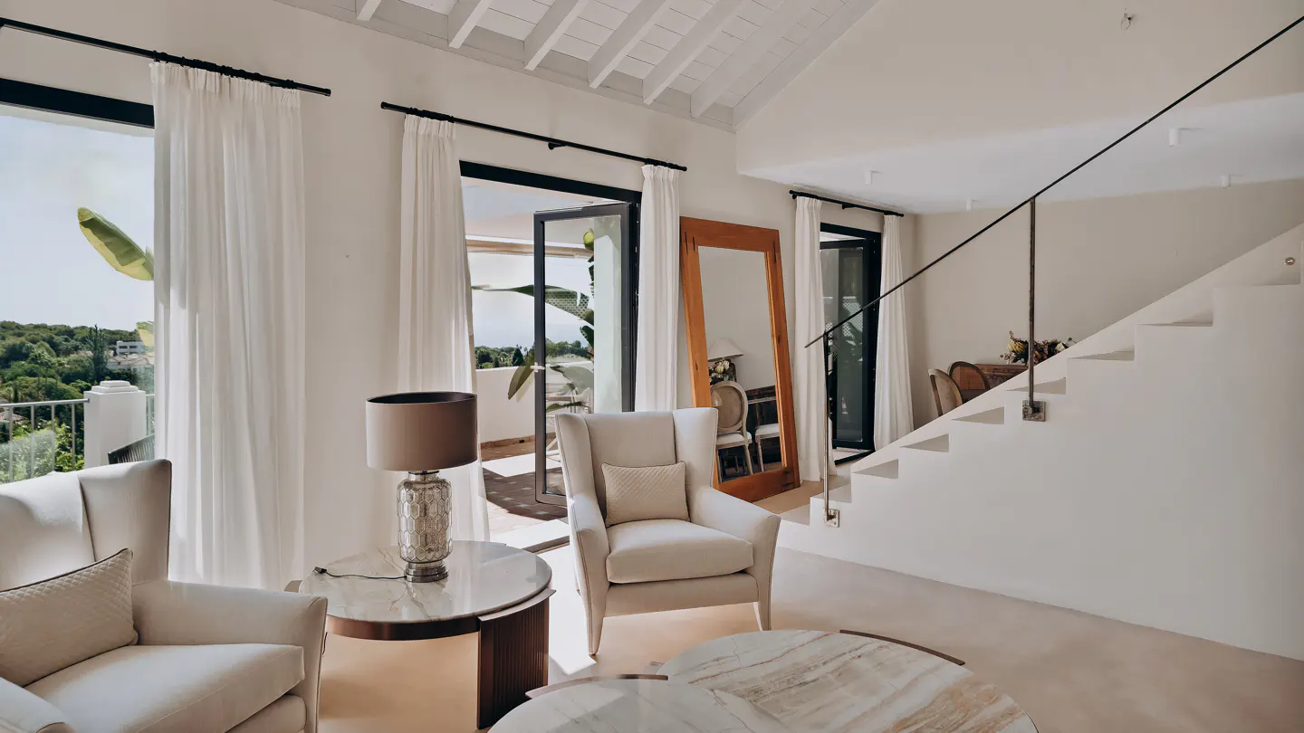 Bright living room with white walls, chairs, and curtains. A staircase with a black railing leads up. Balcony with green trees visible through open doors.