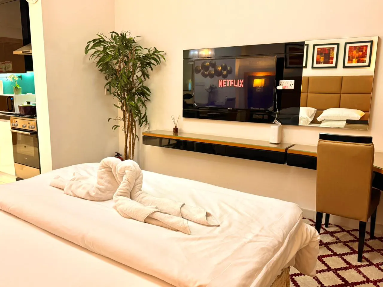 Modern hotel room with a white bed, swan towel art, TV with Netflix, desk, and kitchenette in the background.