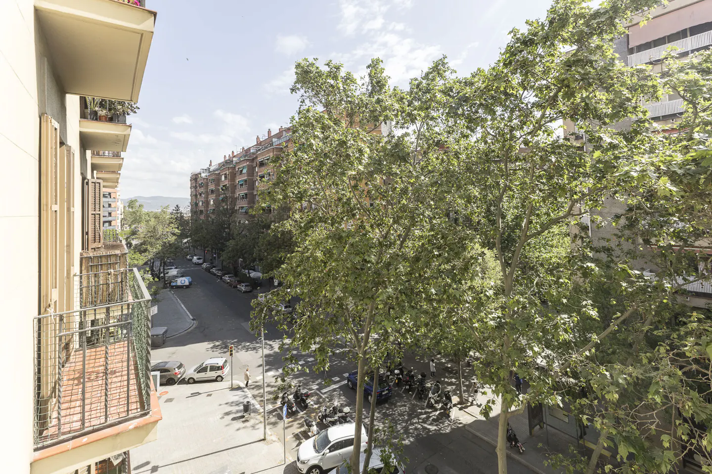View from a balcony overlooking a city street with cars, trees, and apartment buildings on a sunny day.