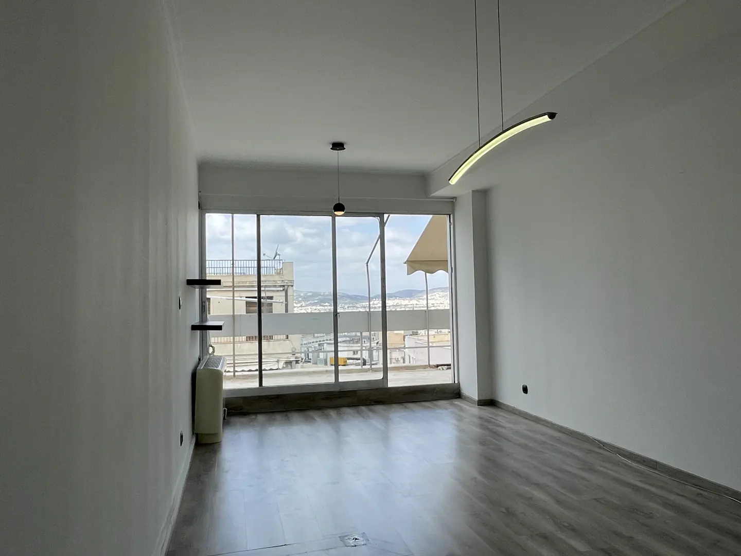 Bright, empty room with wood floors and white walls. Large windows offer a city view. Modern lighting fixtures hang from the ceiling.