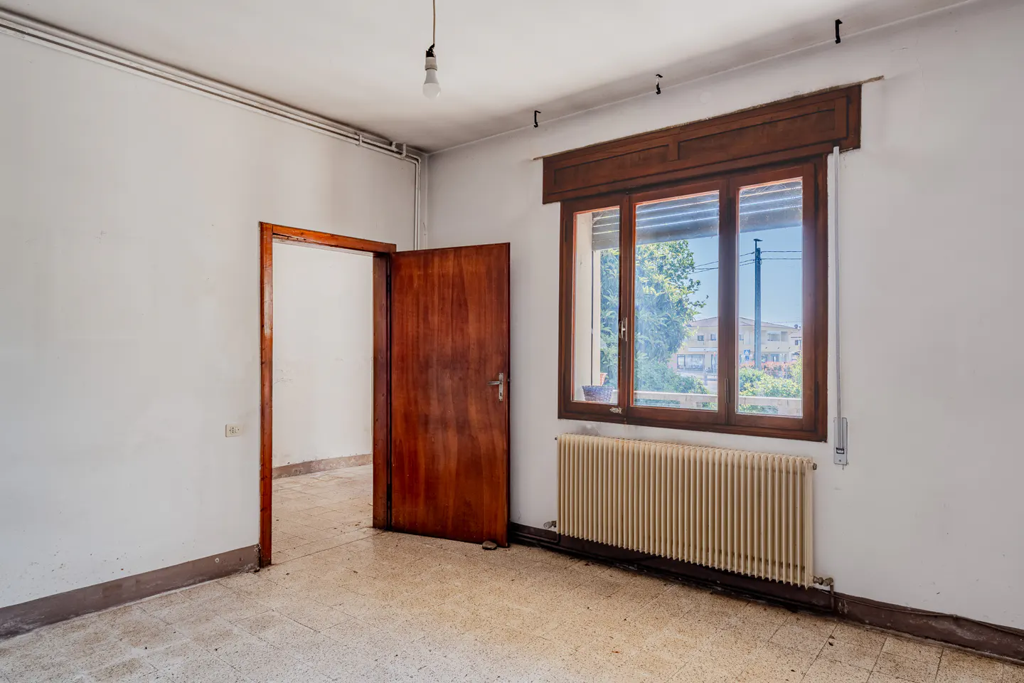 A vacant room with white walls, a brown door ajar, and a window with a brown frame. A radiator sits beneath the window.
